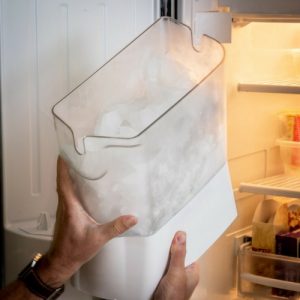 Hands holding an ice container being removed from a refrigerator, illustrating the ice maker installation process.