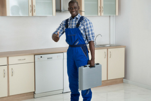 Smiling plumber in blue overalls giving a thumbs-up while holding a toolbox in a modern kitchen, representing customer service in plumbing and heating.