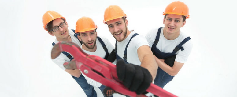 Four young plumbers in safety helmets holding a large wrench, smiling and posing for the camera, emphasizing teamwork in the plumbing profession.