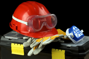 Red hard hat, safety goggles, work gloves, and a respirator on a toolbox, emphasizing essential protective gear for plumbing and heating professionals.