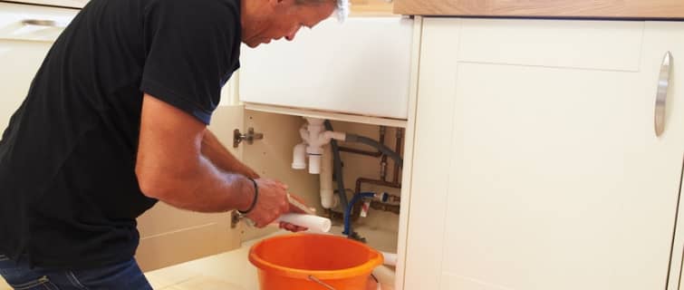 Man attempting DIY plumbing repair under a kitchen sink, using a pipe wrench and working near an orange bucket, illustrating common plumbing mistakes made by amateur plumbers.