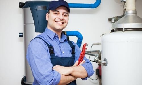 Plumber smiling with adjustable wrench in front of water heater and plumbing system, representing Force Plumbing and Heating LLC's water heater services and maintenance expertise.