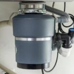 Gray garbage disposal unit with plumbing pipes and electrical connections, showcasing practical kitchen appliance efficiency.