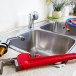 Dual stainless steel kitchen sink with plumbing tools including a red pipe wrench and flexible hoses for maintenance.