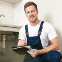 Smiling male plumber in overalls holds a clipboard next to a sink, representing Force Plumbing and Heating LLC.