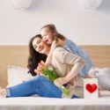 Mother and daughter in a casual indoor setting, holding flowers and a heart-embellished gift box, showcasing their bond.