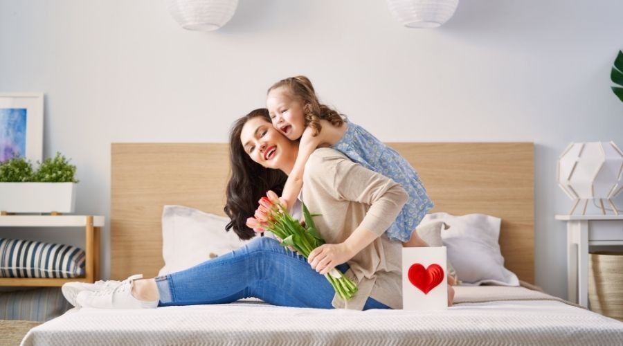 Mother and daughter in a bright room, holding a tulip bouquet and heart card, promoting family warmth and care.