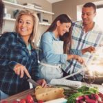 Two women and one man cooking in a bright kitchen with fresh vegetables and utensils for 4th of July celebrations.