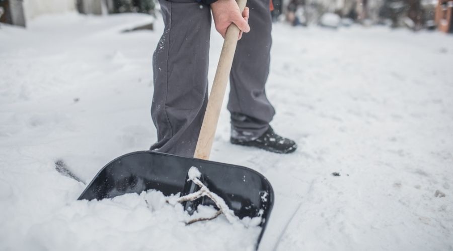 Person shoveling snow on a property, using a black shovel with a wooden handle and wearing black boots.