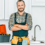 Man in work attire with a tool belt, displaying plumbing tools in a bright kitchen for Force Plumbing and Heating LLC.