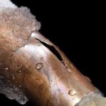 Close-up of a metal pipe showing a crack, ice layer, and water droplets; highlights plumbing risks during winter.