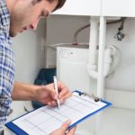 A man performing maintenance checks with a clipboard near plumbing fixtures under a sink for Force Plumbing and Heating LLC.