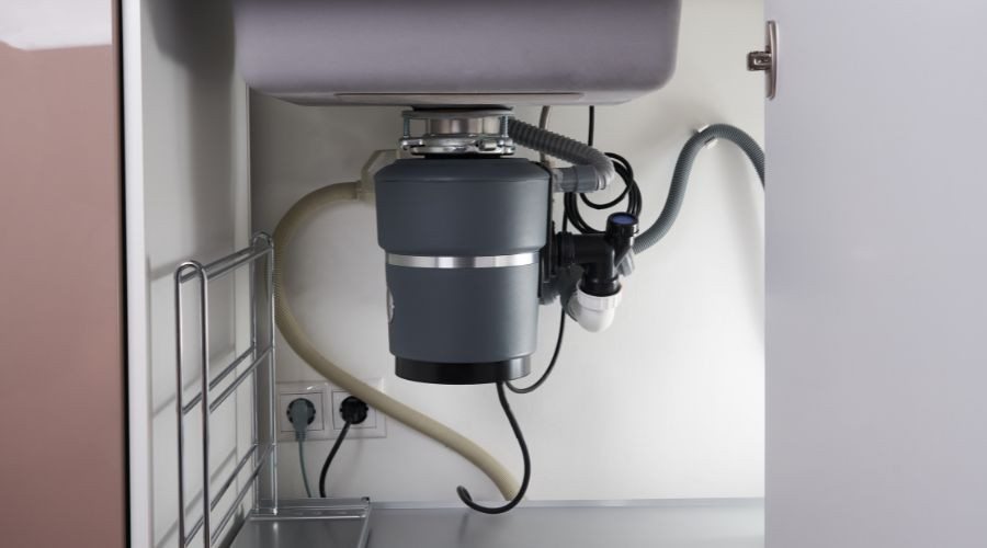 Gray garbage disposal unit under sink with plumbing connections and outlet, showcasing efficient plumbing solutions.